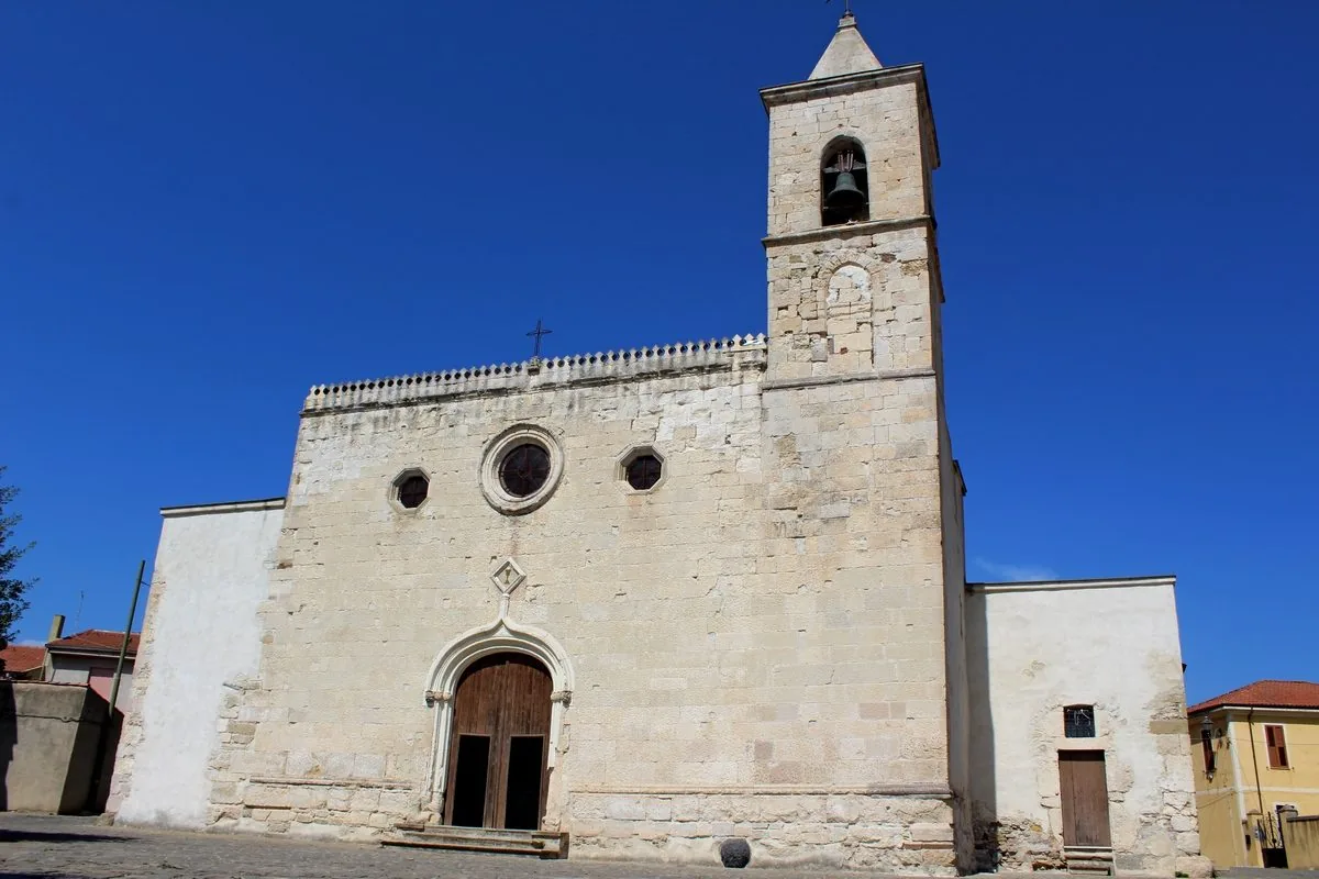 Facciata in pietra chiara della chiesa di San Vito Martire a Gergei, con portale ad arco acuto, rosone e campanile a destra sotto un cielo blu