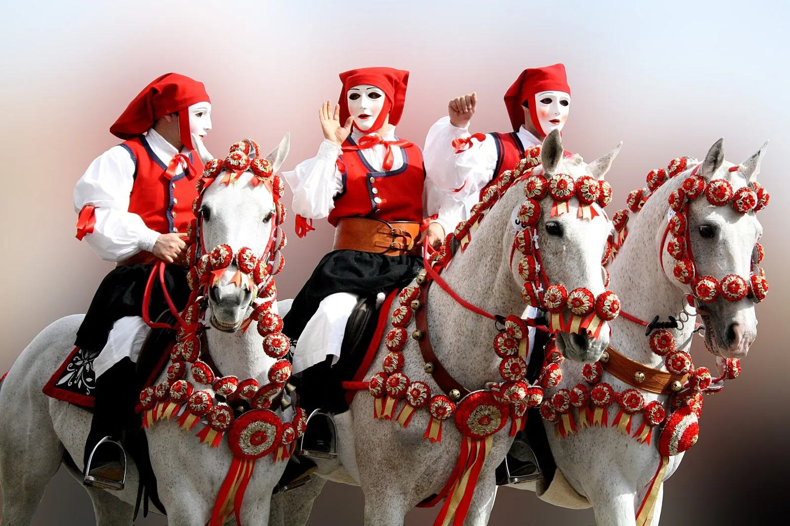 Tre cavalieri della Sartiglia con maschere bianche, berretti rossi e gilet rossi su cavalli grigi bardati di coccarde rosse e oro