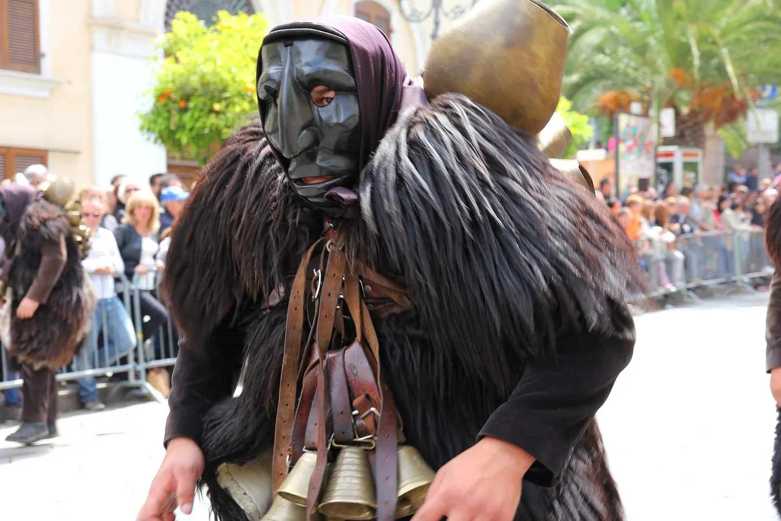 Un Mamuthone con maschera nera di legno, pelli di pecora scure e campanacci sul petto e sulla schiena durante una sfilata, con il pubblico sullo sfondo