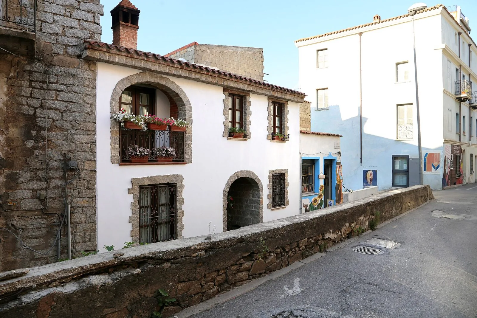 Casa tradizionale di Orgosolo con muri bianchi, archi e cornici in pietra e un balcone fiorito, accanto a edifici decorati con murales
