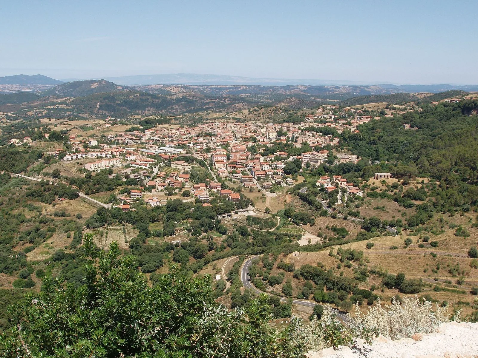 Veduta dall'alto di un paese dai tetti rossi adagiato tra colline boscose, con una strada tortuosa in primo piano e rilievi all'orizzonte