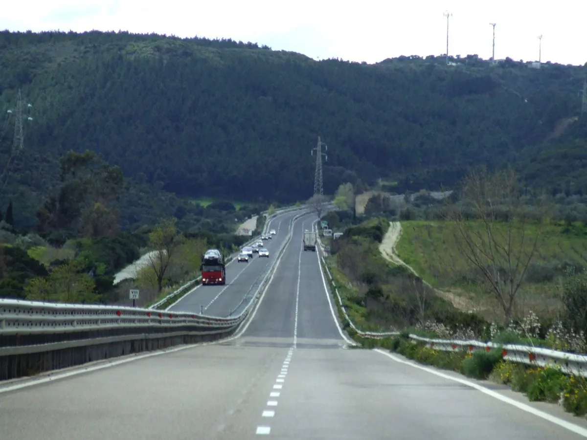 Superstrada a quattro corsie che corre tra colline ricoperte di bosco in Sardegna, con alcune auto e un camion in lontananza