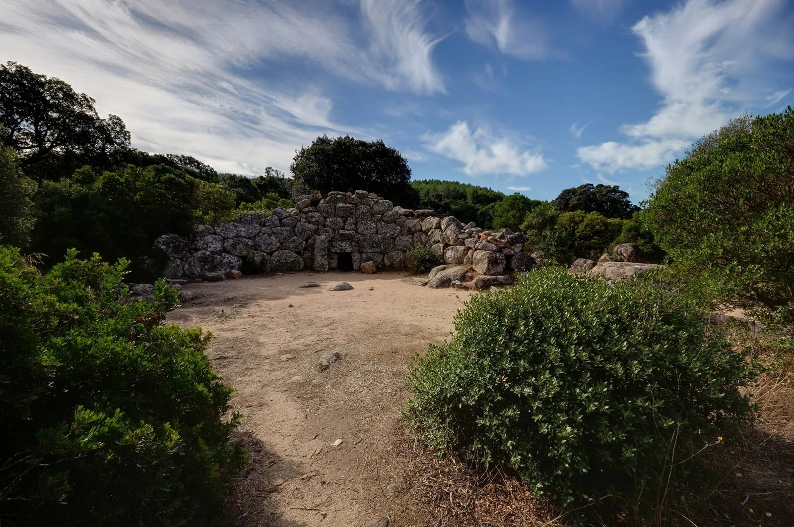 Tomba dei giganti in blocchi di granito con piccolo ingresso centrale, circondata da macchia mediterranea sotto un cielo azzurro con nuvole